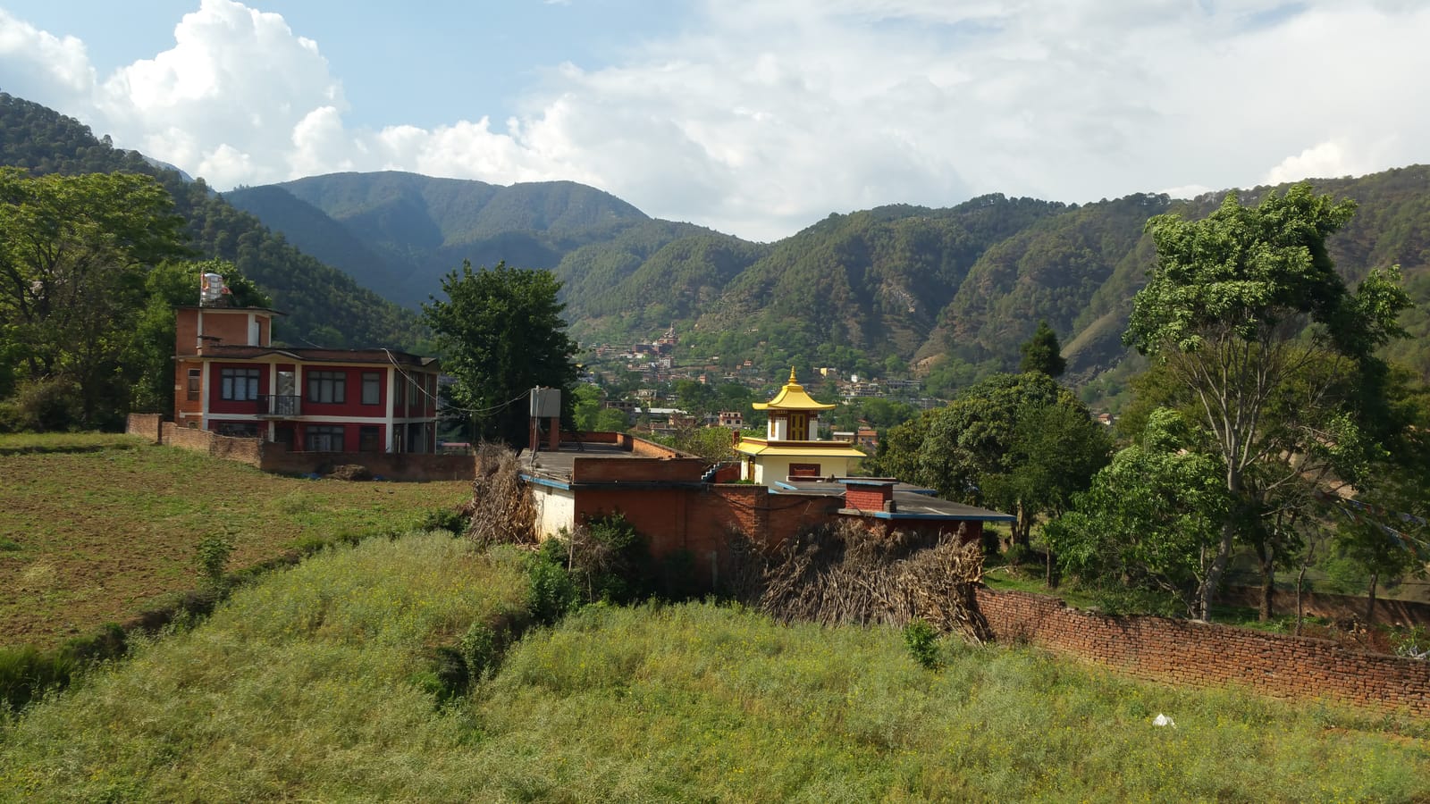 A green hillside village in a Nepalese valley, with a golden-roofed temple among trees and forested mountains beyond